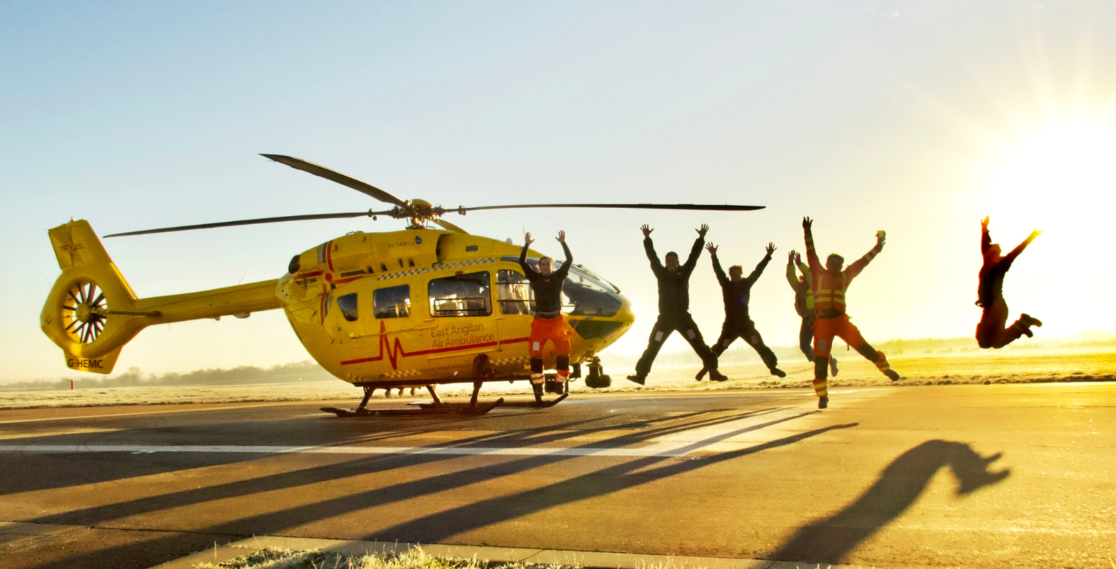 Crew jumping next to yellow, EAAA helicopter at sunset creating shadows on the ground