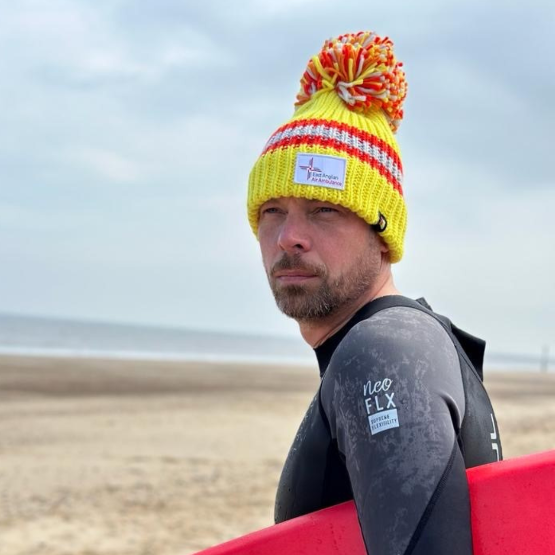 Man wearing yellow and Orange East Anglian Air Ambulance branded bobble hat on a beach in Norfolk